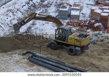 An excavator digs a trench in winter at a construction site. Bricks, concrete slabs and construction waste are lying on the construction site. A yellow excavator throws sand around the site.