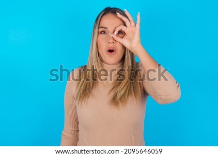 Young hispanic girl wearing turtleneck over blue background  doing ok gesture shocked with surprised face, eye looking through fingers. Unbelieving expression.
