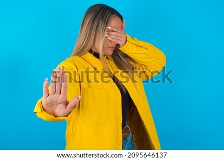 Young hispanic girl wearing yellow waterproof raincoat outerwear over blue background covers eyes with palm and doing stop gesture, tries to hide from everybody.