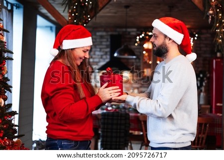 middle aged man and woman in Santa hat stand near Christmas tree in chalet living room decorated for celebrating New Year and Christmas and give each other wrapped present or gift,real life and people