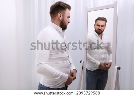 Upset man wearing tight shirt in front of mirror at home. Overweight problem Royalty-Free Stock Photo #2095729588
