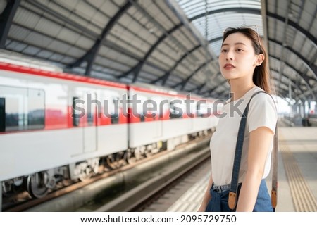 Selective focus half-body image of a beautiful Asian female traveler standing waiting for an arrival of skytrain on a platform with a blurred train in opposite side as a background at skytrain station Royalty-Free Stock Photo #2095729750