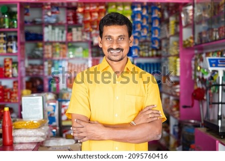 Portrait of successful Indian kirana or groceries businessman standing confidently with smile by looking at camera - concept of small business entrepreneur. Royalty-Free Stock Photo #2095760416