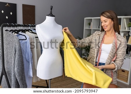 Female fashion designer works on new womenswear collection for clients in cozy workshop studio, dressmaker, tailor or needlewoman standing near clothing rack with fashionable stylish handmade clothes