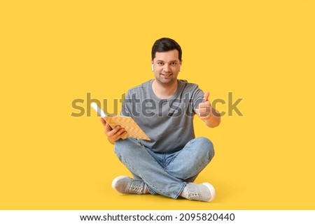 Young man with book and earphones showing thumb-up on color background
