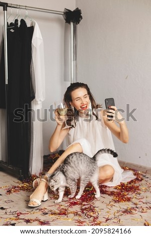 Attractive woman sitting in evening white event dress and celebrating her birthday, Christmas or new year. Holiday concept, happy woman, wine glass with champagne, make selfie, cat. Focus on phone.