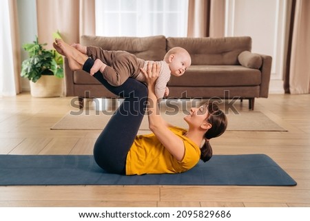New mom lifting her adorable little baby with her legs, doing post-natal fitness exercise. Sports mother is engaged in fitness and yoga with a baby at home. Healthy motherhood and postpartum recovery Royalty-Free Stock Photo #2095829686
