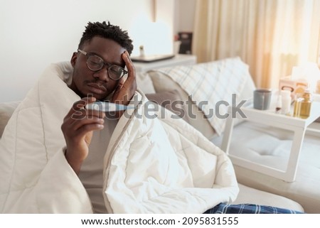 Sick man, wrapped in a duvet, is measuring a temperature while sitting on the bed. Tensed young African-American man with hand on head looking at thermometer while lying in bed at home during the day.