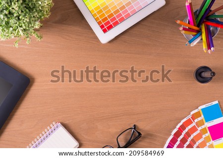 Modern office workplace with digital tablet, notepad, colorful pencils, glasses, graphic tablet, stylus, cup of coffee, and color swatches on a desktop. View from the top