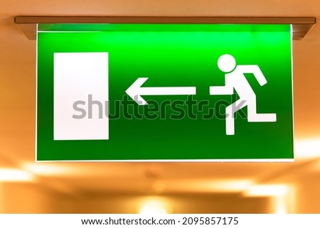 A bright green emergency evacuation sign with a directional arrow and a running figure hanging from the ceiling in the hallway. Selective focus. Close-up