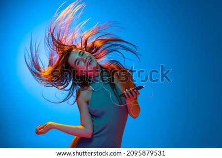 Dancing and listening music. Young happy beautiful girl with long hair isolated on blue studio backgroud in neon light. Human emotions, facial expression concept. Fashion, style, beauty and ad