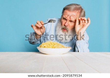 Fastidious senior hairy gray-bearded man tasting large portion of noodles, pasta isolated on blue studio background. Food, festival, ad, taste and dieting, humor. Healthy and unhealthy eating