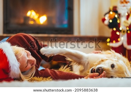 A beautiful woman lying down on the floor at home with her Retriever at Christmas holidays. She is wearing a Santa Claus hat and she is looking at the camera. Family dog Christmas tree concept