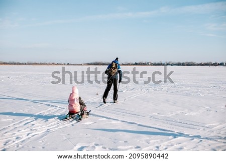 Family on winter walk. Young man and child are skiing in winter on frozen river near forest, man is sledding child in snow.  Royalty-Free Stock Photo #2095890442