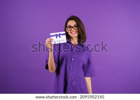Young woman in a shirt on a purple background with a gift certificate smiling cheerfully, with makeup, lipstick on her lips and glasses, excited