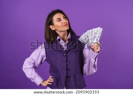 Young woman in a shirt and vest on a purple background with a bundle of dollars of money in her hand smiles confidently, looks thoughtfully to the right