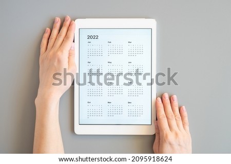 tablet computer with an open app of calendar for 2022 year in a womans hands on a gray background. concept business or to do list goals with technology using. top view, flat lay
