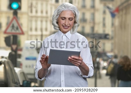 Smiling senior lady using digital tablet standing on the city street.