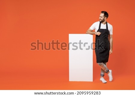 Full length young man barista bartender barman employee in apron t-shirt work in coffee shop leaning on blank board promotional content place text isolated on orange background Small business startup Royalty-Free Stock Photo #2095959283