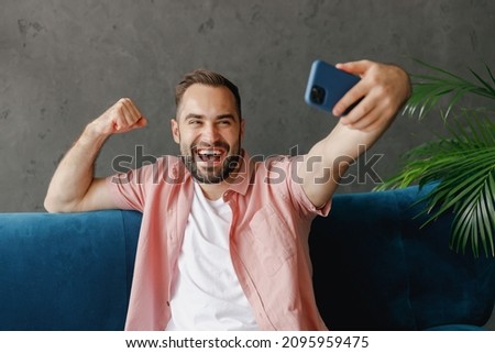 Young happy man in casual clothes do selfie shot on mobile cell phone post photo social network talk video call do winner gesture sit on blue sofa at home flat indoors rest relax on weekends free time
