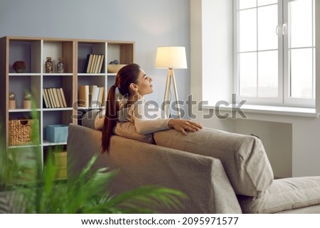 Woman uses chance to relax, unwind and get some peace of mind on comfy couch at home. Happy lady enjoying silence and quiet leisure time in beautiful room with comfortable sofa and Nordic wooden shelf Royalty-Free Stock Photo #2095971577