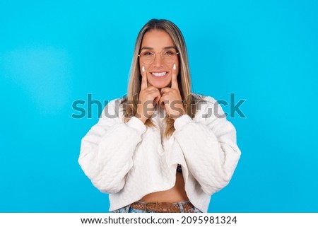 Happy Young hispanic girl wearing white knitted sweater over blue background with toothy smile, keeps index fingers near mouth, fingers pointing and forcing cheerful smile