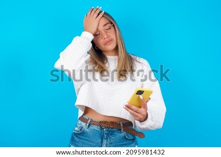 Upset depressed Young hispanic girl wearing white knitted sweater over blue background makes face palm as forgot about something important holds mobile phone expresses sorrow and regret blames