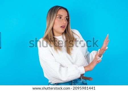 Displeased Young hispanic girl wearing white knitted sweater over blue background keeps hands towards empty space and asks not come closer sees something unpleasant