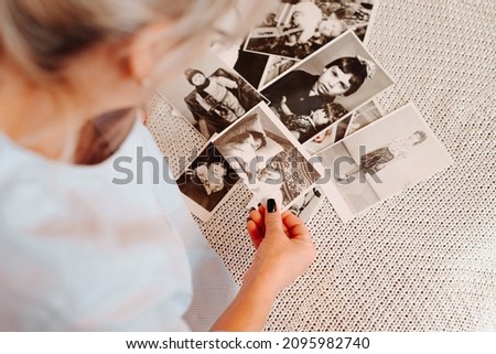 Close-up caucasian woman looking retro photographs of childhood and youth, nostalgic sitting on sofa at home. Back view of female hands holding black and white film photos. Selective focus on pictures Royalty-Free Stock Photo #2095982740