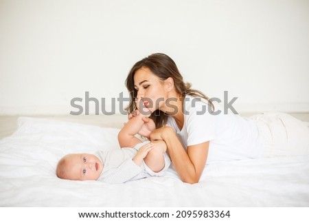 mom kisses babys legs at home in the bedroom. Happy young mom hugs her little daughter in bed, top view. Mother and daughter are resting in bed at home. Good morning!