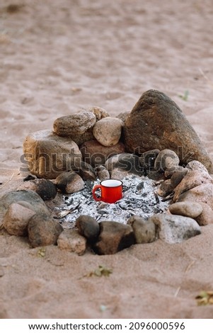 hiking and outdoor activities, a beautiful campfire and a red cup of coffee or tea. picnic by the sea or lake on a sandy beach with large stones. camping life and hiking or wilderness tourism. burn