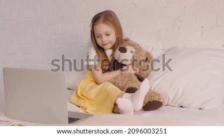 Child hugging teddy bear while looking at laptop in bedroom
