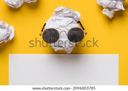 Crumpled paper and sunglasses on it look like a face, next to a white sheet for text, on a yellow background. Copy space. Top view. 