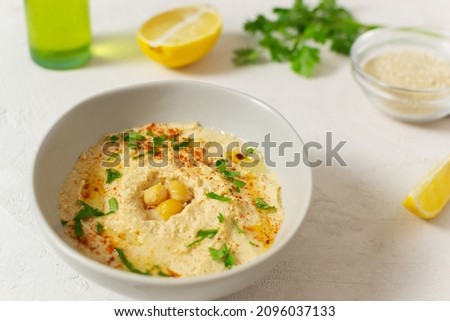 Hummus in a ceramic white bowl with paprika and parsley leaves and ingredients on a light background. Banner. A traditional Middle Eastern dish. Horizontal orientation. Vegetarian food concept.