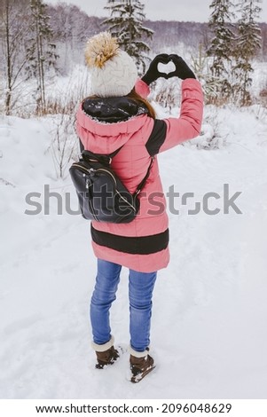 Winter forest and woman walking in snow nature. Rear view of girl in pink color down jacket and warm hat strolls in woods. Hands in gloves shows heart