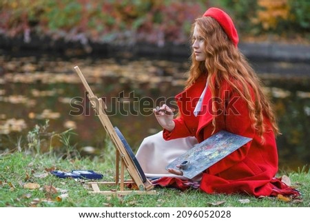 girl-artist, in a red coat and beret, sits on the grass, in the fall and paints a picture. Art, creative people, crises, inspiration