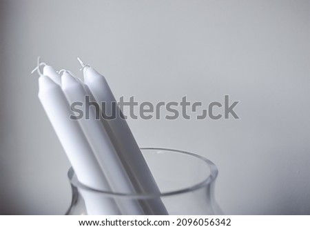 Many tall white candles with white wicks in a glass vase standing on a wooden handmade stool against minimal wall background. DIY Christmas holiday decor. Copy space. 
