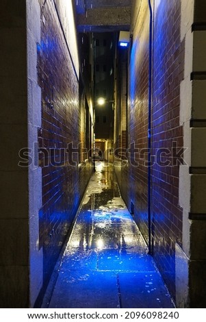 A narrow London alleyway in the rain, with a blue coloured light reflecting off the ground and walls.  Image has copy space.