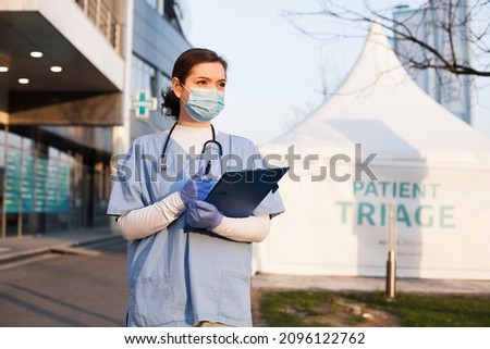Young beautiful caucasian female NHS doctor in front of emergency ambulance clinic,next to white patient admission triage tent for receiving infected Coronavirus patients,COVID-19 quarantine hospital Royalty-Free Stock Photo #2096122762