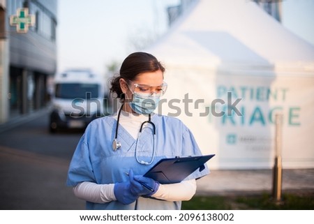 Female medical NHS doctor checking patient admission clipboard form in front of hospital,frontline first responder next to patient triage tent with ambulance car in background,first aid rescue service Royalty-Free Stock Photo #2096138002