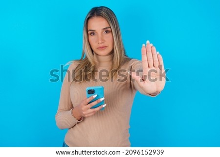 Young hispanic girl wearing turtleneck over blue background  using and texting with smartphone with open hand doing stop sign with serious and confident expression, defense gesture