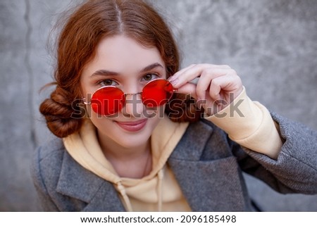 Portrarit of fashion young red hair woman wearing red glasses and casual style clothes, posing against street wall