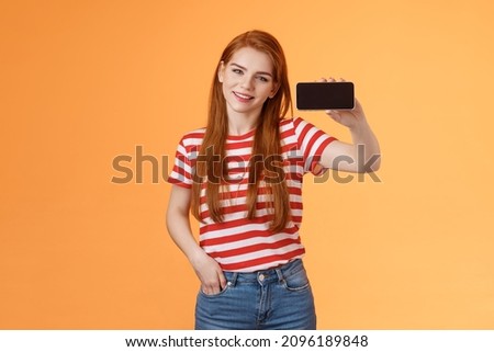 Sassy confident attractive 20s caucasian ginger girl introduce smartphone app, recommend cool game, show cellphone screen horizontal, smiling proud, brag beating score, stand orange background