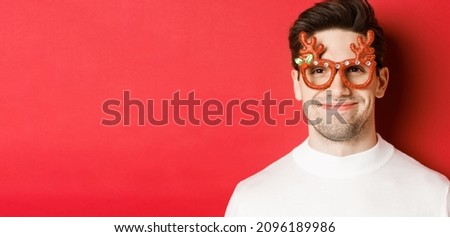 Concept of winter holidays, christmas and celebration. Close-up of attractive smiling male model in party glasses, standing against red background