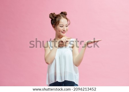 Lady holding palm up and pointing to her right while looking at you smiling in pink background