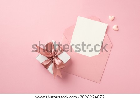 Top view photo of white giftbox with pink satin ribbon bow small hearts and open pink envelope with paper card on isolated pastel pink background with blank space