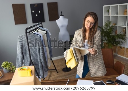Female Fashion Designer In Studio Working On Sketches Or Documents At Desk With Laptop. No time to relax. Young woman tailor is standing near drapery and modeling new clothes in design studio
