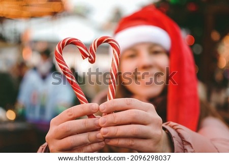 In the background, a blurry young happy smiling woman in a Santa hat holding in the foreground a heart symbol made of red and white striped sweets in the form of a cane, express support, love.