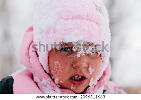 Closeup photo of female kid with sad face covered with snow and about to cry wearing pink winter clothes in forest. Astonishing background full of white color and snow.  Royalty-Free Stock Photo #2096311663