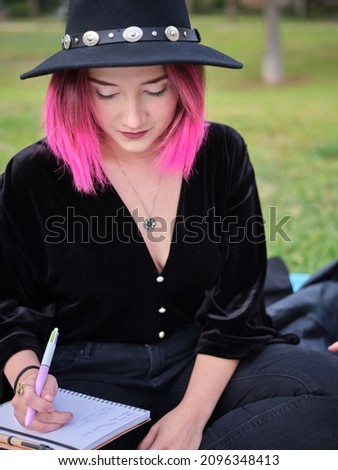 portrait of a young girl with pink hair and a black hat sitting with a notebook